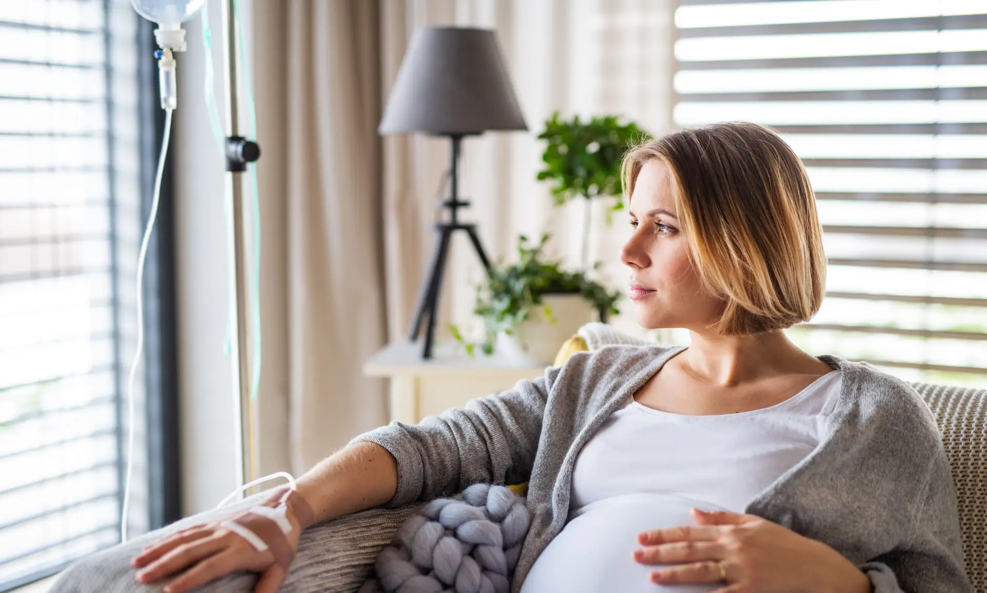 Woman receiving in-home IV therapy while seated on a couch, looking thoughtfully out a window, with IV drip setup visible.
