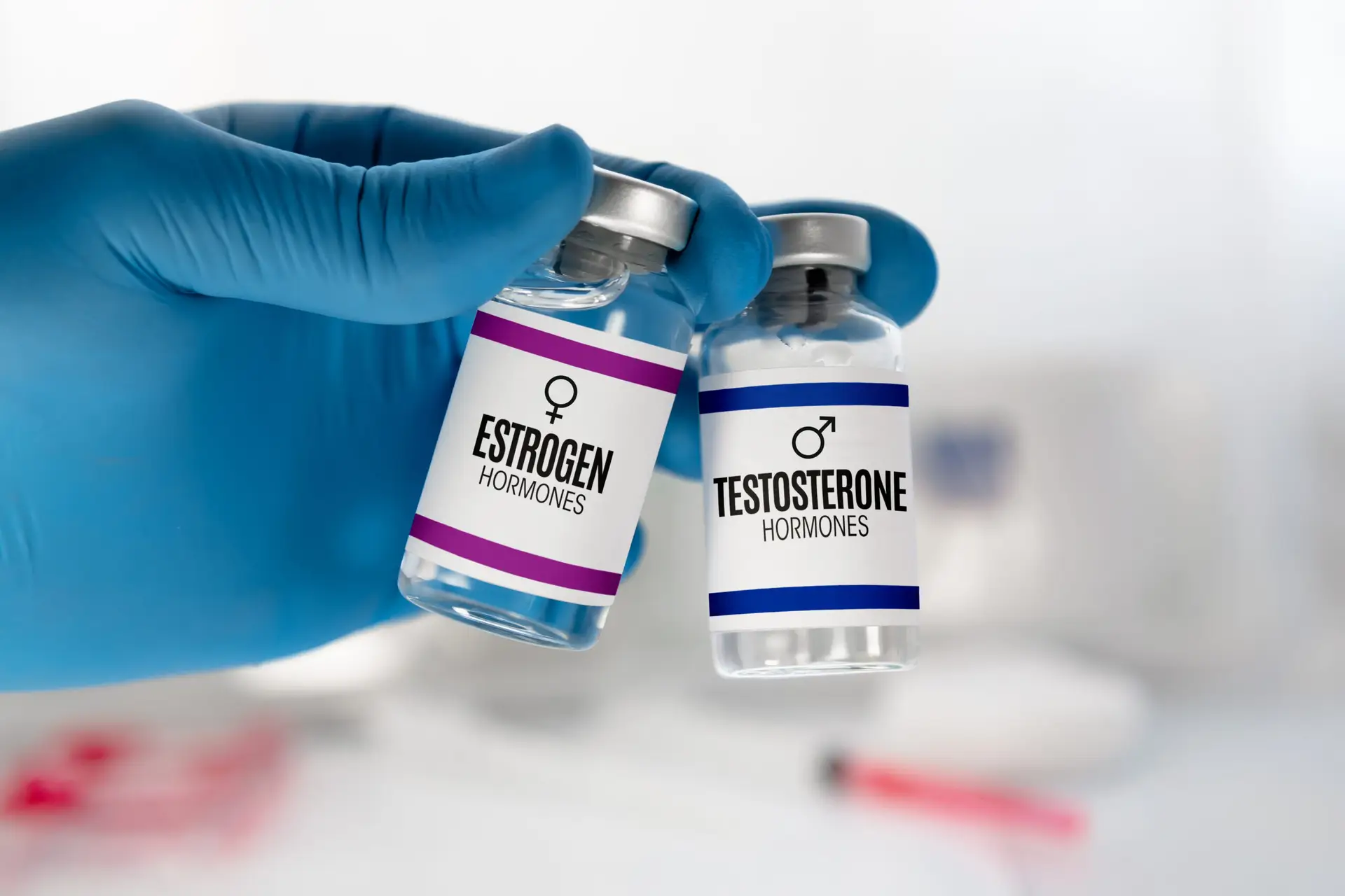 Gloved hand holding vials labeled "Estrogen" and "Testosterone," representing bio-identical hormone replacement therapy (HRT) for personalized health solutions.
