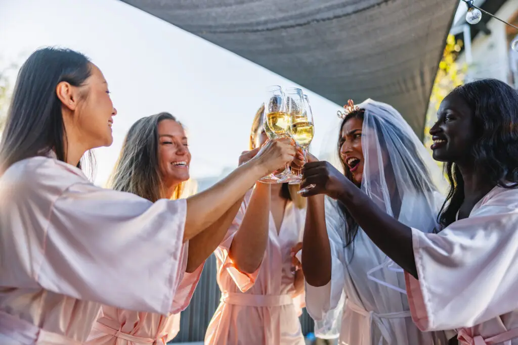 Group of women in matching robes celebrating with champagne glasses, including a bride-to-be in a veil, capturing the joy of bachelorette party festivities.